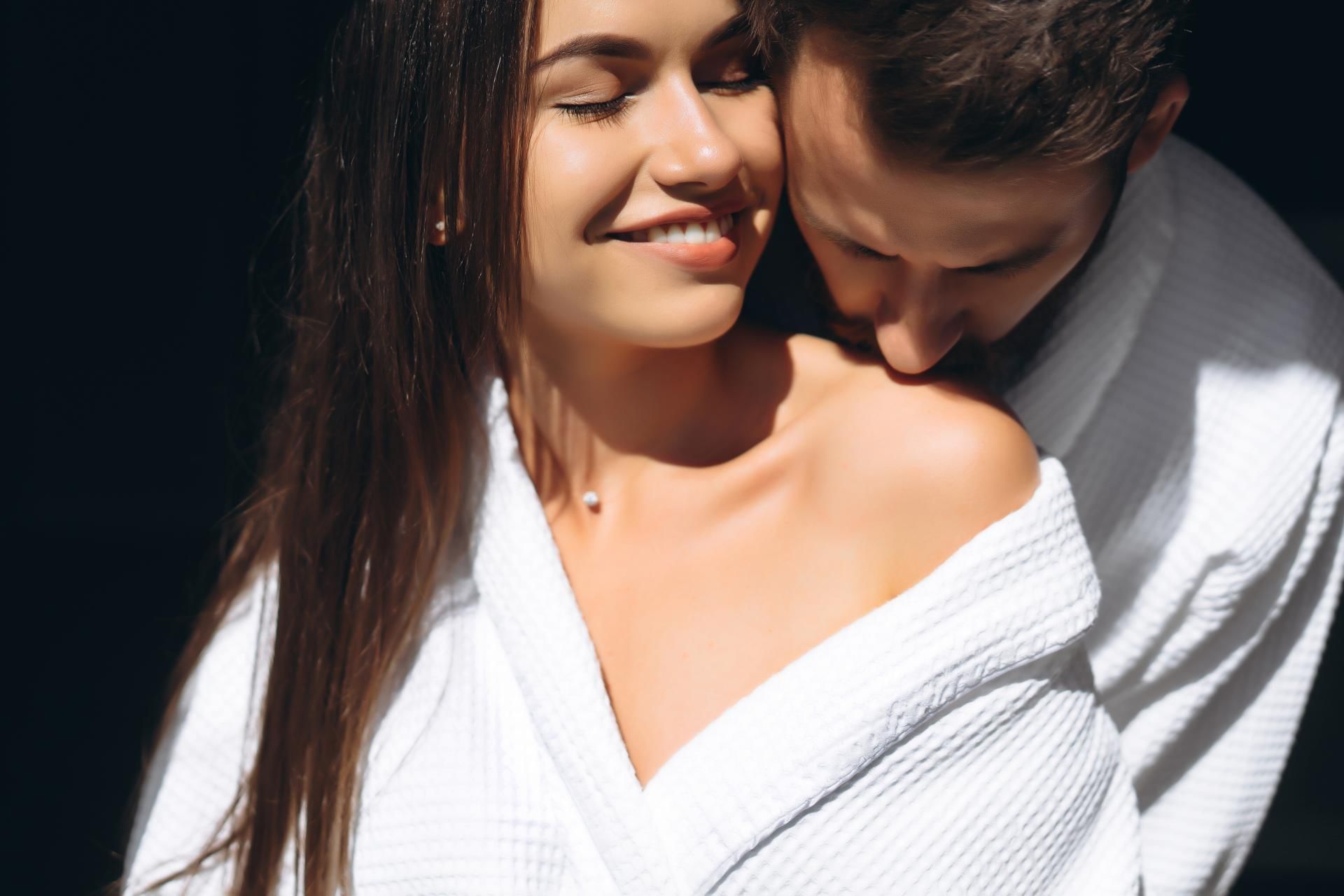 Pareja abrazada, ambos con batas blancas, ella sonríe con ojos cerrados, él la mira de cerca.